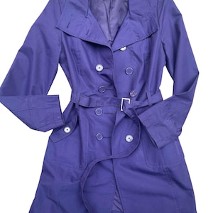 May include: A double-breasted, purple trench coat with a matching belt. The coat features a notched collar, button closures, and pockets. The belt has a silver-colored buckle. The coat is made of a smooth fabric.