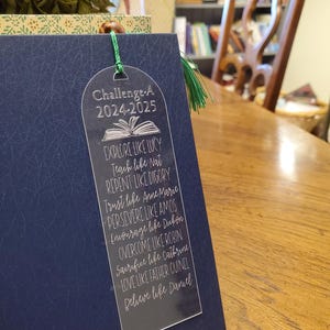 Pode incluir: Um marcador de livros de acr&iacute;lico transparente com um borla verde, colocado em um livro azul marinho. O marcador de livros diz "Challenge A 2024-2025" e inclui frases inspiradoras como "Explore como Lucy" e "Acredite como Daniel."