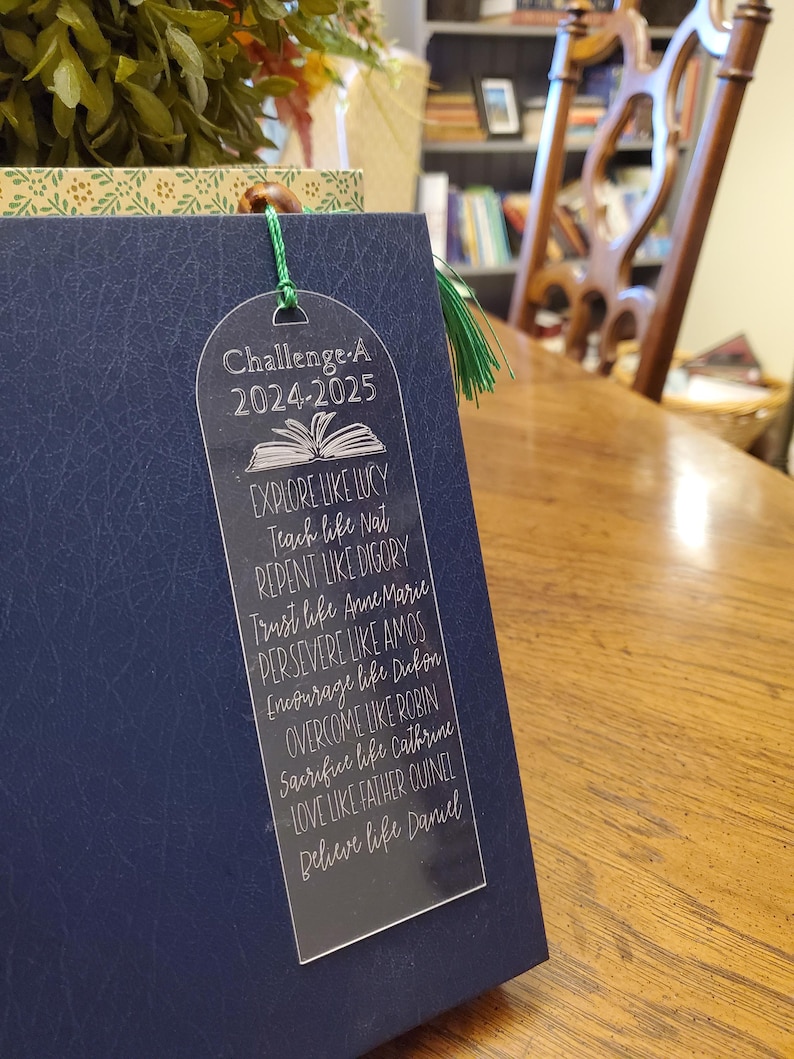 Pode incluir: Um marcador de livros de acr&iacute;lico transparente com um borla verde, colocado em um livro azul marinho. O marcador de livros diz "Challenge A 2024-2025" e inclui frases inspiradoras como "Explore como Lucy" e "Acredite como Daniel."