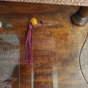 May include: A clear acrylic bookmark with a pink tassel and a wood bead. The bookmark features the text "Proverbs 25:2" and the quote "It is the glory of God to conceal a thing, but the glory of kings is to search things out".