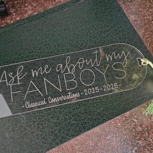 May include: A clear acrylic bookmark with the text "Ask me about my FANBOYS - Classical Conversations - 2025-2026" in white script. The bookmark is on a dark green leather-bound book and has a green tassel and wooden bead attached.