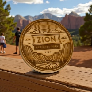 Puede incluir: Placa redonda de madera con la inscripción "ZION NATIONAL PARK". El diseño incluye un paisaje de montañas, un río y estrellas. La placa es de color marrón claro, probablemente un recuerdo o un objeto decorativo.