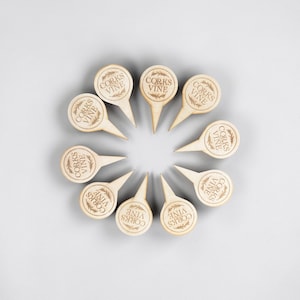 May include: A top-down view of ten wooden wine charms arranged in a circle. Each charm has a circular top with the words "CORKS VINE" and a leaf design, and a pointed stem. The charms are light brown against a gray background.