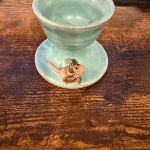 May include: A handmade, turquoise ceramic egg cup with a small, brown and tan mouse design on the base. The cup has a rounded bowl shape and sits on a small, circular base. The ceramic piece is glazed and has a rustic, artisanal feel.