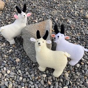 Può includere: Tre peluche di capra bianche all'uncinetto con corna nere e nasi rosa. I peluche sono seduti su un letto di ciottoli grigi.