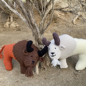 Peut inclure: Deux animaux en peluche crochetés, un bison brun et orange et une chèvre blanche et grise, sont posés l'un à côté de l'autre sur une surface sablonneuse. Le bison a une longue queue et la chèvre a une longue queue et une barbe.