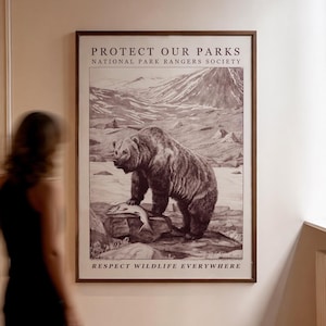May include: A framed sepia-toned print depicting a grizzly bear with a fish, set against a mountain backdrop. The print's text reads "PROTECT OUR PARKS" and "RESPECT WILDLIFE EVERYWHERE". The artwork is displayed on a wall.