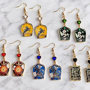 May include: Five pairs of gold-toned dangle earrings featuring enamel charms with various designs, including house crests and the text I Solemnly Swear That I'm Up to No Good, accented with small heart-shaped crystals in yellow, green, red, blue, and black.