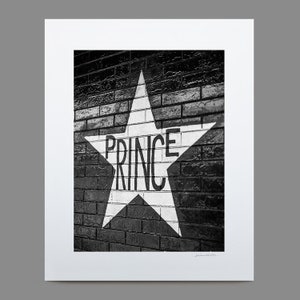 May include: A black and white photograph of a white star painted on a brick wall. The star has the word "PRINCE" written inside of it.