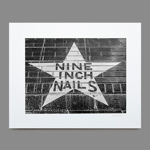 May include: Black and white photograph of a star painted on a brick wall. The star contains the words "NINE INCH NAILS" in a bold, sans-serif font. The image is framed with a white border.