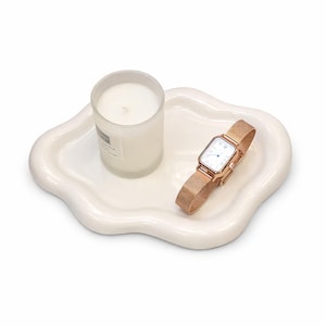 May include: A white, cloud-shaped ceramic tray holds a clear glass candle and a rose gold wristwatch with a mesh band. The watch has a rectangular face with a white dial and black Roman numerals. The candle is cylindrical.