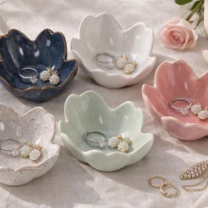 Könnte beinhalten: Eine Sammlung von Schmuckschalen aus Keramik in Blütenblattform und verschiedenen Farben: Blau, Weiß, Rosa und gesprenkelt. Jede Schale enthält Ringe und Ohrringe. Eine Rose und eine Schmuckschatulle befinden sich im Hintergrund.