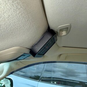 May include: A black electronic device, possibly a remote control, is mounted on the interior ceiling of a car. The device has buttons and a display, and is positioned near the sun visor. The car's interior is beige.