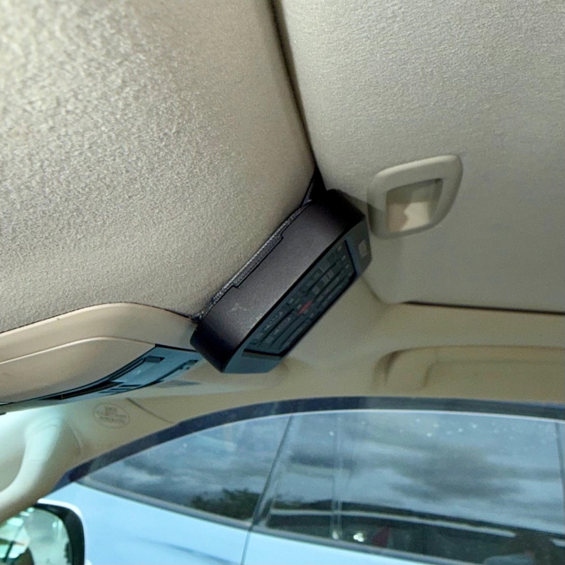May include: A black electronic device, possibly a remote control, is mounted on the interior ceiling of a car. The device has buttons and a display, and is positioned near the sun visor. The car's interior is beige.