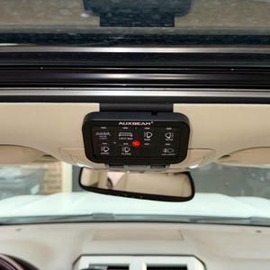 May include: A black AUXBEAM light control panel with illuminated icons for roof light, light bar, side, rear, hood, flood, and fog lights. The panel is mounted on a vehicle's interior ceiling, above the rearview mirror.