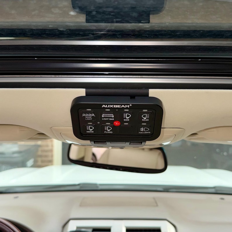 May include: A black AUXBEAM light control panel with illuminated icons for roof light, light bar, side, rear, hood, flood, and fog lights. The panel is mounted on a vehicle's interior ceiling, above the rearview mirror.