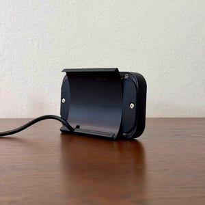 May include: A black rectangular electronic device with a hinged cover and a curved black cable extending from the side. The device is on a wooden surface, with a neutral-coloured wall in the background.