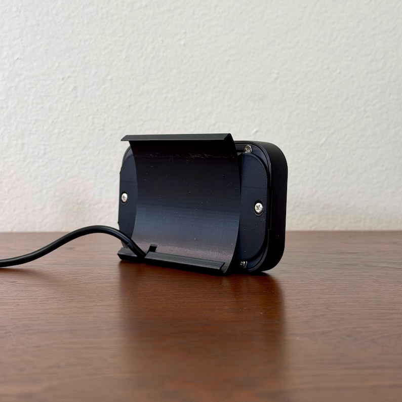 May include: A black rectangular electronic device with a hinged cover and a curved black cable extending from the side. The device is on a wooden surface, with a neutral-coloured wall in the background.