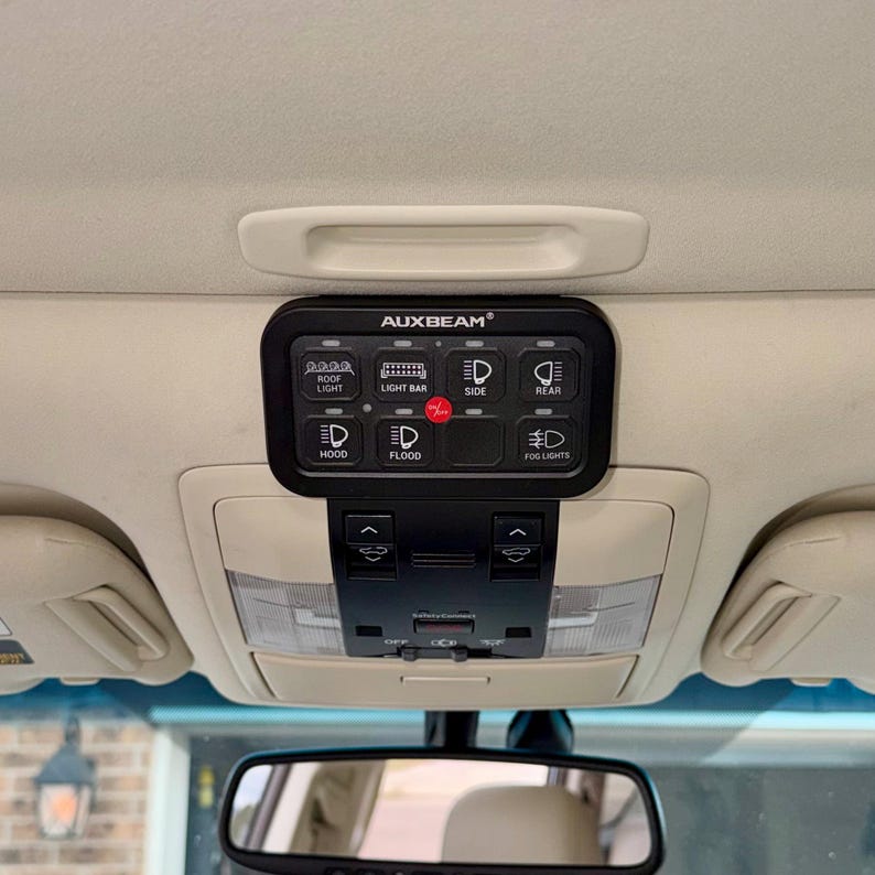 May include: Black AUXBEAM light control panel with various labelled buttons for roof light, light bar, side, rear, hood, flood, and fog lights. The panel is mounted on a vehicle's ceiling, above the rearview mirror.
