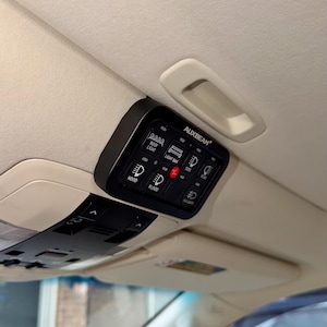 Op de afbeelding: Zwart AUXBEAM-schakelaar paneel met verlichte pictogrammen voor daklicht, lichtbalk, motorkap, floodlight, zijkant, achterkant en mistlampen. Het paneel is gemonteerd op het interieurplafond van een voertuig.