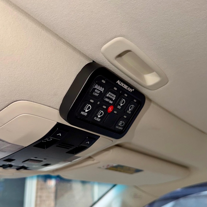 May include: Black AUXBEAM switch panel with illuminated icons for roof light, light bar, hood, flood, side, rear, and fog lights. The panel is mounted on a vehicle's interior ceiling, near a white grab handle.