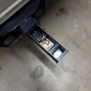 Puede incluir: Una caja de almacenamiento metálica negra montada en el enganche, parcialmente abierta, que revela un llavero de coche, llaves, monedas, un billete doblado, un objeto cilíndrico y un disco blanco. La caja está instalada en el receptor de enganche de un vehículo.