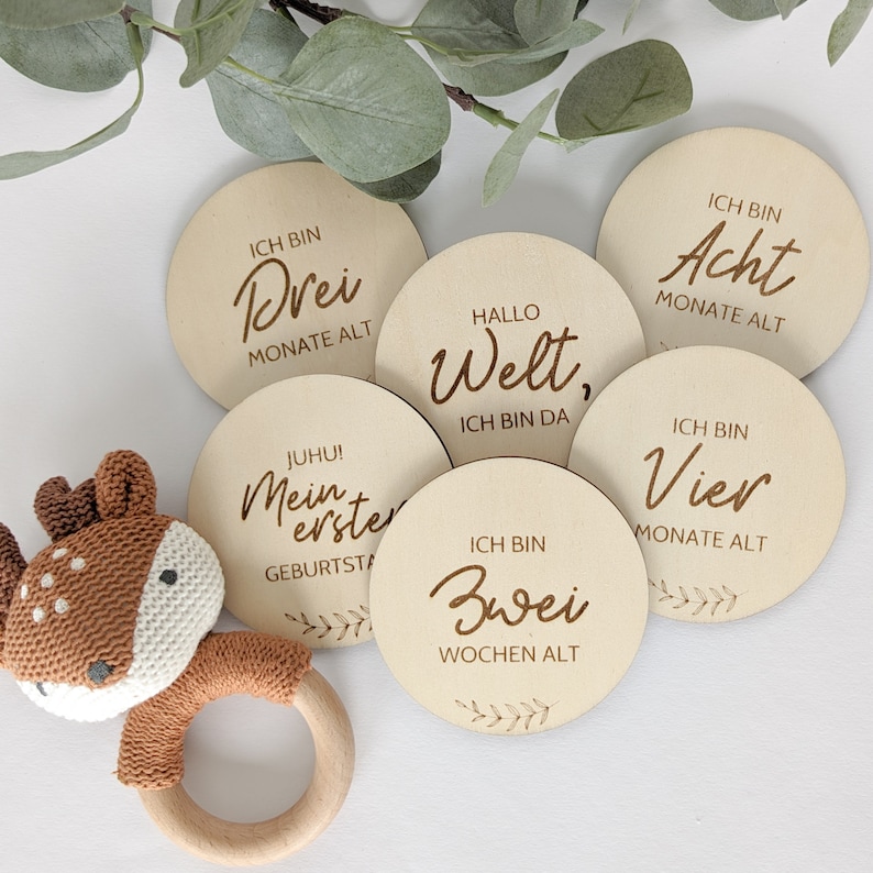 May include: Wooden baby milestone cards with phrases in German, including "Ich bin Drei Monate alt" (I am three months old), "Ich bin Acht Monate alt" (I am eight months old), "Ich bin Vier Monate alt" (I am four months old), "Hallo Welt, Ich bin da" (Hello world, I am here), "Juhu! Mein erster Geburtstag" (Hooray! My first birthday), and "Ich bin Zwei Wochen alt" (I am two weeks old).