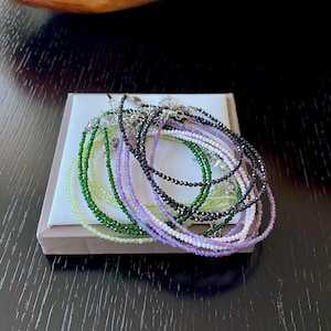 Peut inclure: Plusieurs bracelets de perles délicats de différentes couleurs, dont vert, violet, noir et blanc. Les bracelets sont présentés sur une petite plateforme carrée blanche.
