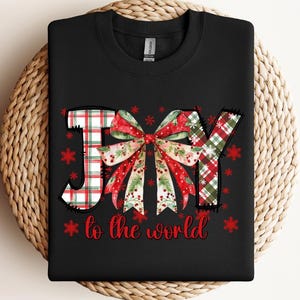 May include: A black crewneck sweatshirt featuring a graphic design with the word JOY, where the letter O is replaced by a large red, green, and white plaid bow. The text to the world is printed in red below, surrounded by small red snowflakes.