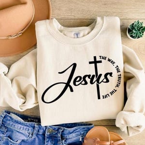 Könnte beinhalten: Cremefarbenes Sweatshirt mit dem Wort "Jesus" in Schreibschrift, begleitet von einem Kreuz und dem Text "The Way, The Truth, The Life" in Schwarz. Das Sweatshirt wird mit blauen Jeansshorts, einem beigen Hut und Sandalen kombiniert.