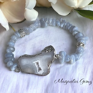 May include: A light blue gemstone bracelet featuring a large, irregularly shaped, silver-edged agate stone as a focal point. The bracelet is composed of small, rounded blue stones and silver accents. The brand name "Magnolia Gemz" is written in cursive.