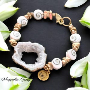 May include: A handcrafted bracelet featuring white spiral beads, gold-toned accents, and a geode pendant. The bracelet includes a gold clasp and a small charm with a floral design. The brand name "Magnolia Gemz" is visible.
