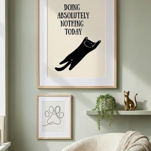 May include: A framed wall art print featuring a black cat silhouette stretching with the text DOING ABSOLUTELY NOTHING TODAY in black letters on a cream background, paired with a smaller framed print showing a single black line drawing of a paw print.