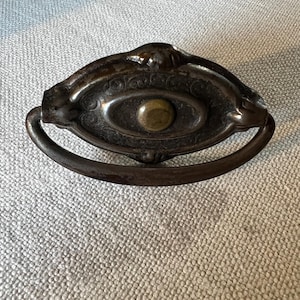 May include: Antique metal drawer pull with an oval center and a curved handle. The center features an ornate design with a brass-colored knob. The pull has a dark, aged finish, suggesting it is vintage or a reproduction.