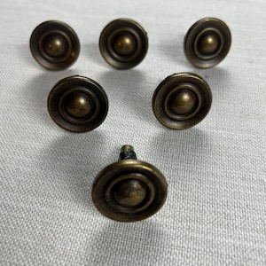 May include: Six antique brass cabinet knobs. Each knob has a circular design with a raised center and concentric rings. The knobs are arranged on a light gray textured surface.