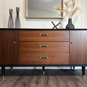 May include: A mid-century modern style credenza with a black frame and legs. The credenza has two doors and three drawers with gold-toned hardware. The wood has a rich, dark brown finish. Decorative items sit on top of the credenza.
