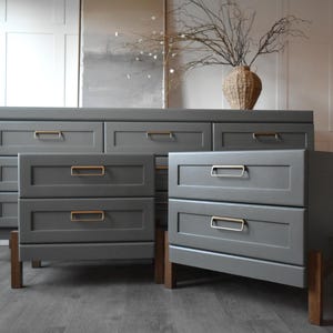 May include: A gray dresser and two matching nightstands with gold-colored drawer pulls. The furniture has a modern design with wooden legs. A woven vase with branches sits on top of the dresser.