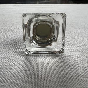 May include: A clear, square-shaped glass object with a central, circular design. The glass has a textured appearance and a slightly tinted interior. The object is likely a decorative element or a functional piece, such as a knob or a stopper.