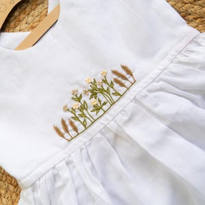 K&ouml;nnte beinhalten: Wei&szlig;es Leinenkleid mit gesticktem Blumen- und Weizendesign. Das Kleid hat einen runden Ausschnitt, Holzkn&ouml;pfe und einen gerafften Rock. Die Stickerei umfasst gr&uuml;ne Bl&auml;tter, kleine wei&szlig;e Bl&uuml;ten und beige Weizen&auml;hren.