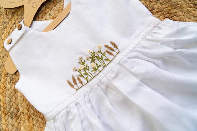 K&ouml;nnte beinhalten: Wei&szlig;es Leinenkleid mit gesticktem Blumen- und Weizendesign. Das Kleid hat einen runden Ausschnitt, Holzkn&ouml;pfe und einen gerafften Rock. Die Stickerei umfasst gr&uuml;ne Bl&auml;tter, kleine wei&szlig;e Bl&uuml;ten und beige Weizen&auml;hren.
