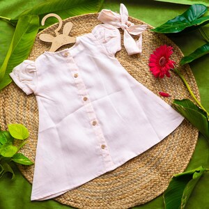 May include: A light pink button-down dress with short, puffed sleeves and wooden buttons. A matching headband with a bow sits nearby. The dress is displayed on a bunny-shaped wooden hanger, with a red gerbera daisy and green leaves.