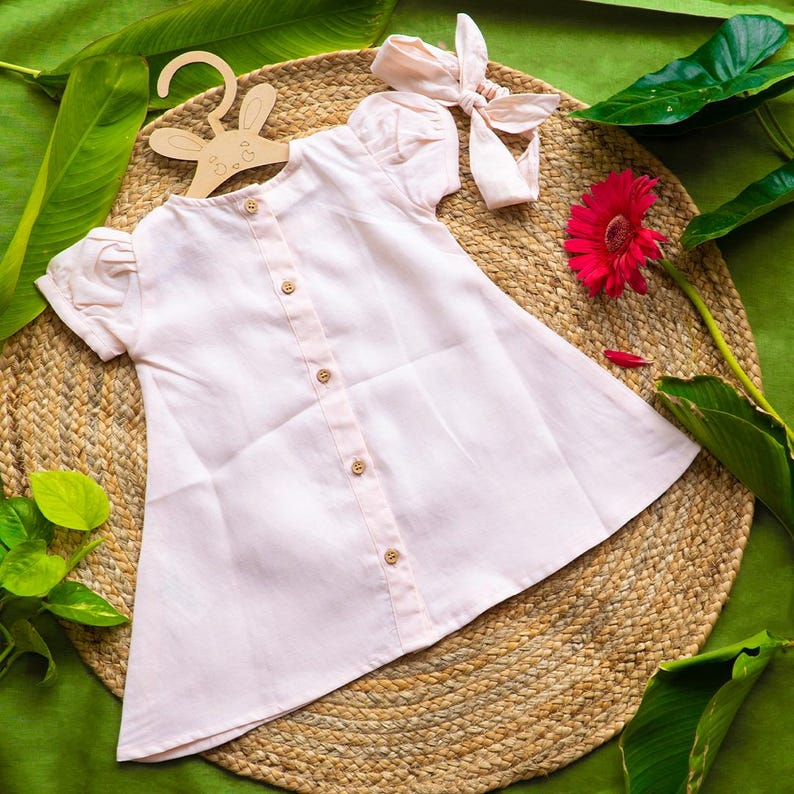 May include: A light pink button-down dress with short, puffed sleeves and wooden buttons. A matching headband with a bow sits nearby. The dress is displayed on a bunny-shaped wooden hanger, with a red gerbera daisy and green leaves.