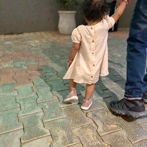 May include: A young child wearing a light pink dress with wooden buttons down the back and pink sneakers. The child is holding an adult's hand while walking on a patterned brick surface.