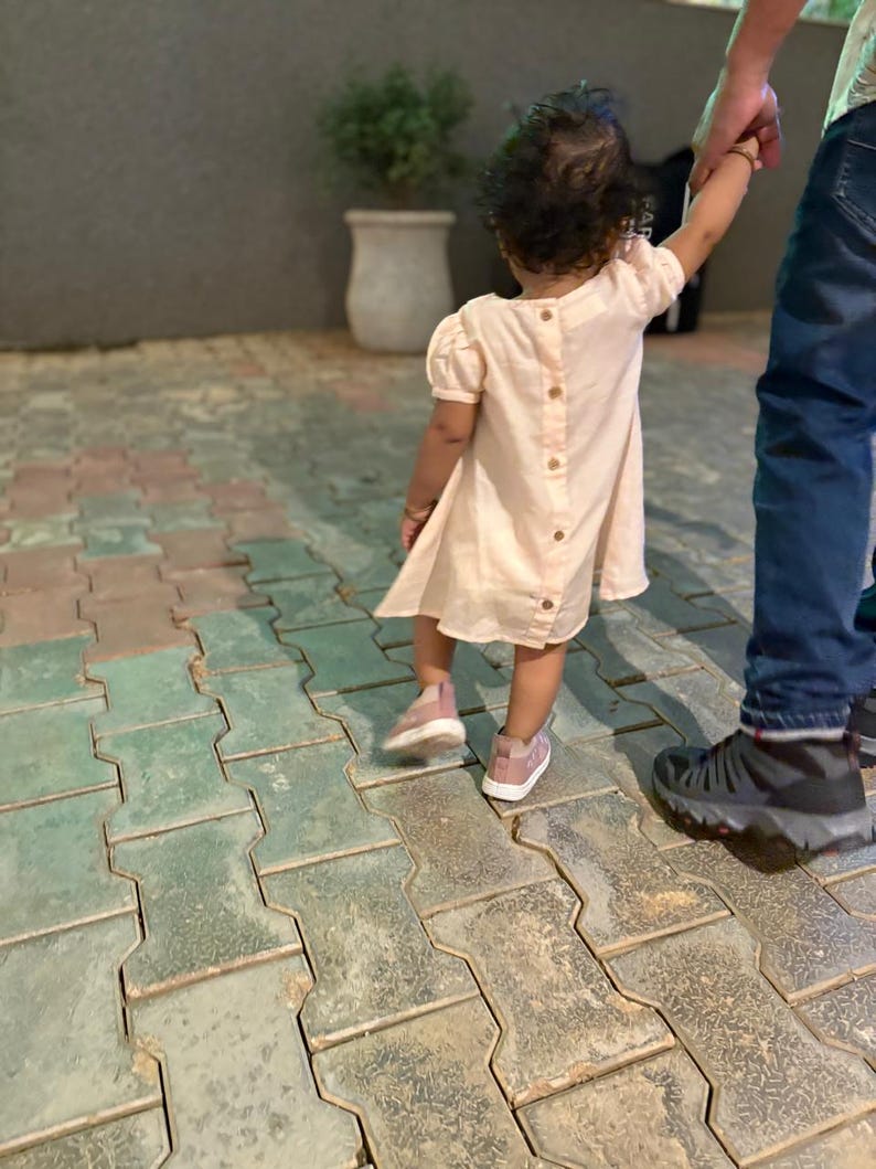 May include: A young child wearing a light pink dress with wooden buttons down the back and pink sneakers. The child is holding an adult's hand while walking on a patterned brick surface.