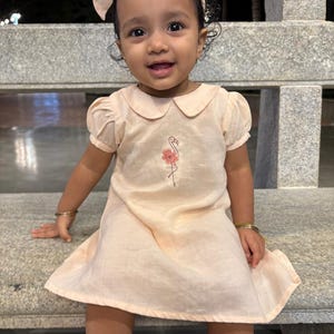 May include: A light peach-colored dress with a flamingo embroidery. The dress has a Peter Pan collar and short, puffed sleeves. A matching bow headband and gold bracelets complete the outfit. The child is sitting on a stone bench.