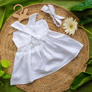 K&ouml;nnte beinhalten: Ein wei&szlig;es Babykleid mit gekreuztem R&uuml;cken, pr&auml;sentiert auf einem h&ouml;lzernen Hasenaufh&auml;nger. Ein passendes wei&szlig;es Stirnband und eine wei&szlig;e Blume liegen neben dem Kleid. Das Kleid ist aus einem weichen, flie&szlig;enden Stoff gefertigt.