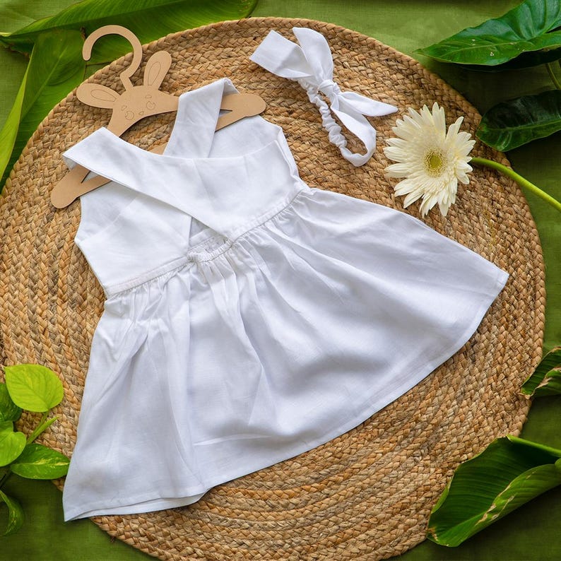 K&ouml;nnte beinhalten: Ein wei&szlig;es Babykleid mit gekreuztem R&uuml;cken, pr&auml;sentiert auf einem h&ouml;lzernen Hasenaufh&auml;nger. Ein passendes wei&szlig;es Stirnband und eine wei&szlig;e Blume liegen neben dem Kleid. Das Kleid ist aus einem weichen, flie&szlig;enden Stoff gefertigt.