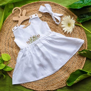 K&ouml;nnte beinhalten: Ein wei&szlig;es &auml;rmelloses Kleid mit floraler Stickerei, pr&auml;sentiert auf einem Hasen-Kleiderb&uuml;gel. Ein passendes wei&szlig;es Stirnband und eine wei&szlig;e Blume sind ebenfalls vorhanden. Das Kleid ist aus leichtem Stoff gefertigt.