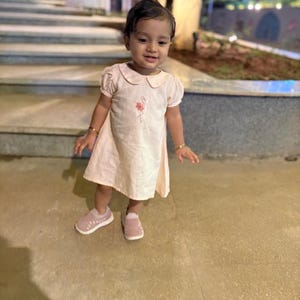 May include: A young child wearing a light pink dress with a Peter Pan collar and short sleeves. The dress features a small floral embroidery. The child is also wearing pink shoes with white soles and gold bracelets.