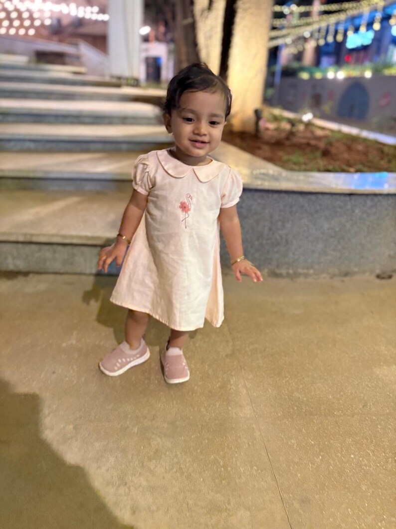 May include: A young child wearing a light pink dress with a Peter Pan collar and short sleeves. The dress features a small floral embroidery. The child is also wearing pink shoes with white soles and gold bracelets.